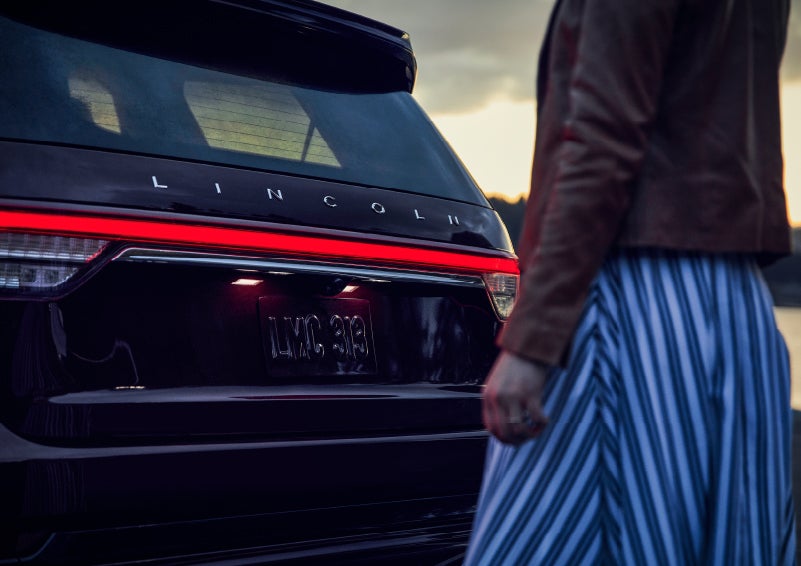 A person is shown near the rear of a 2024 Lincoln Aviator® SUV as the Lincoln Embrace illuminates the rear lights | Nick Mayer Lincoln Mayfield in Mayfield Heights OH