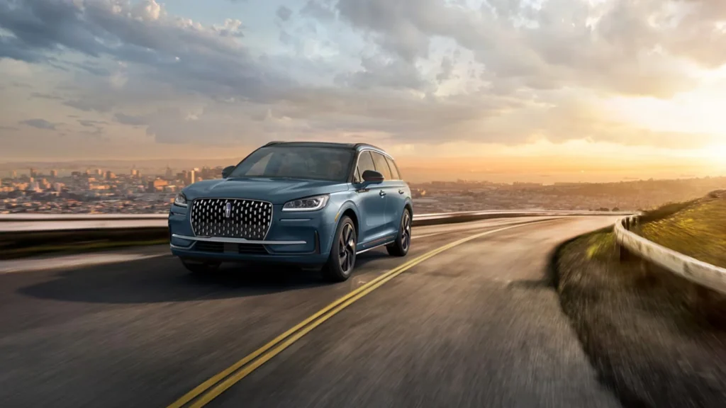 2025 Lincoln Corsair Grand Touring driving along a scenic, winding road at sunset, with a panoramic view of a city skyline in the background and a golden sky above.