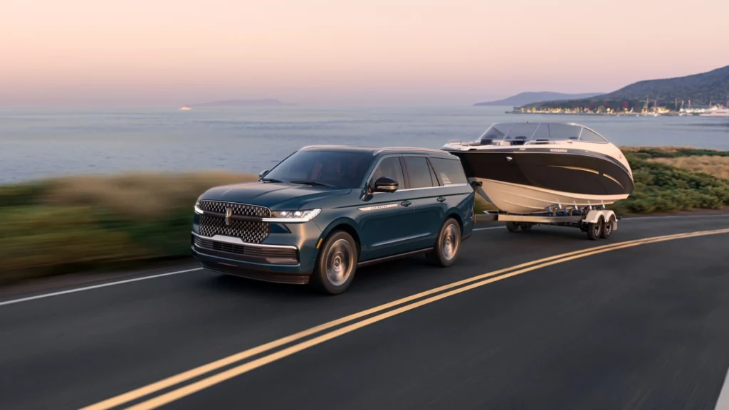 2025 Lincoln Navigator Reserve in a deep teal color towing a speedboat along a coastal highway during sunset, with scenic water and mountain views in the background.