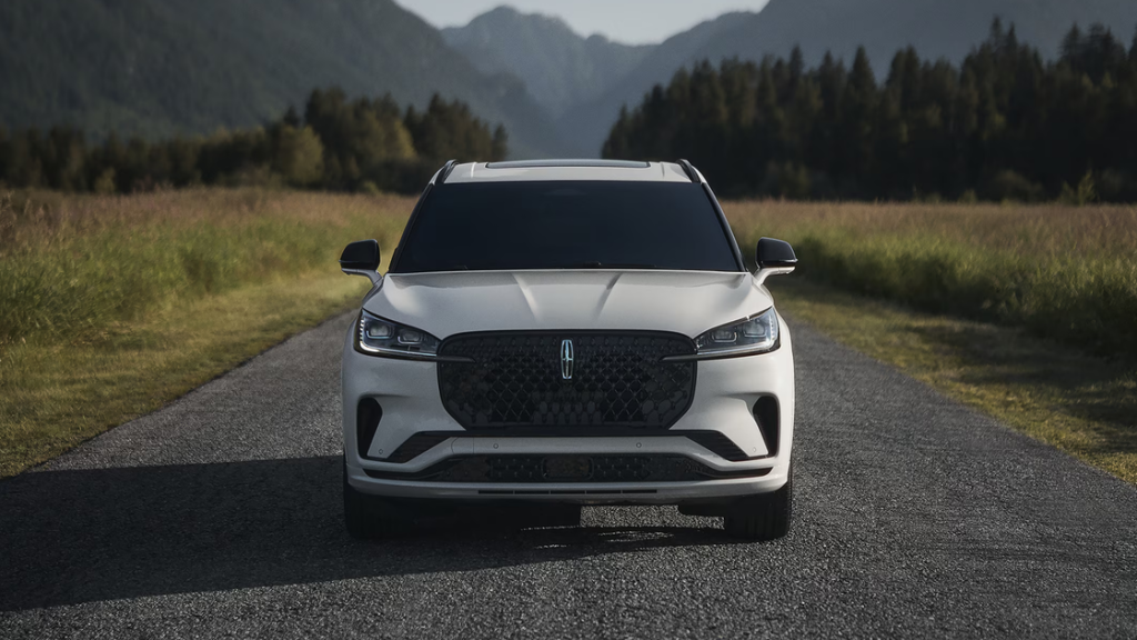 Front view of a white 2025 Lincoln Aviator on a rural road with grassy fields and mountains in the background.