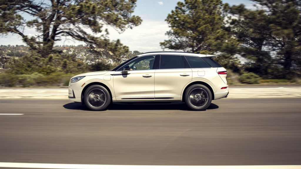 Side view of a white 2025 Lincoln Corsair Grand Touring driving on a suburban road with trees and hills.