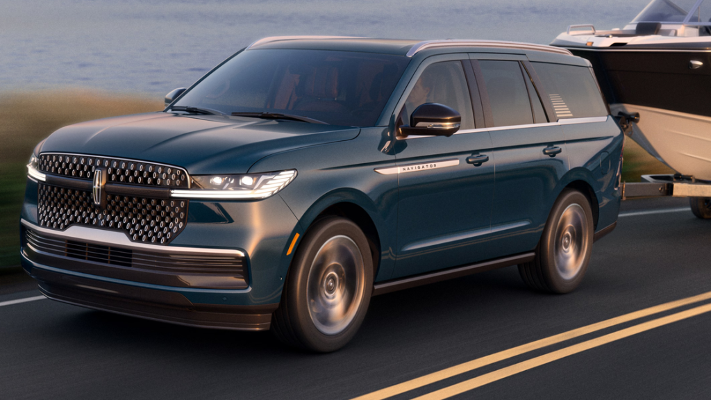 Front-side view of a dark green 2025 Lincoln Navigator towing a boat on a lakeside road at sunset.