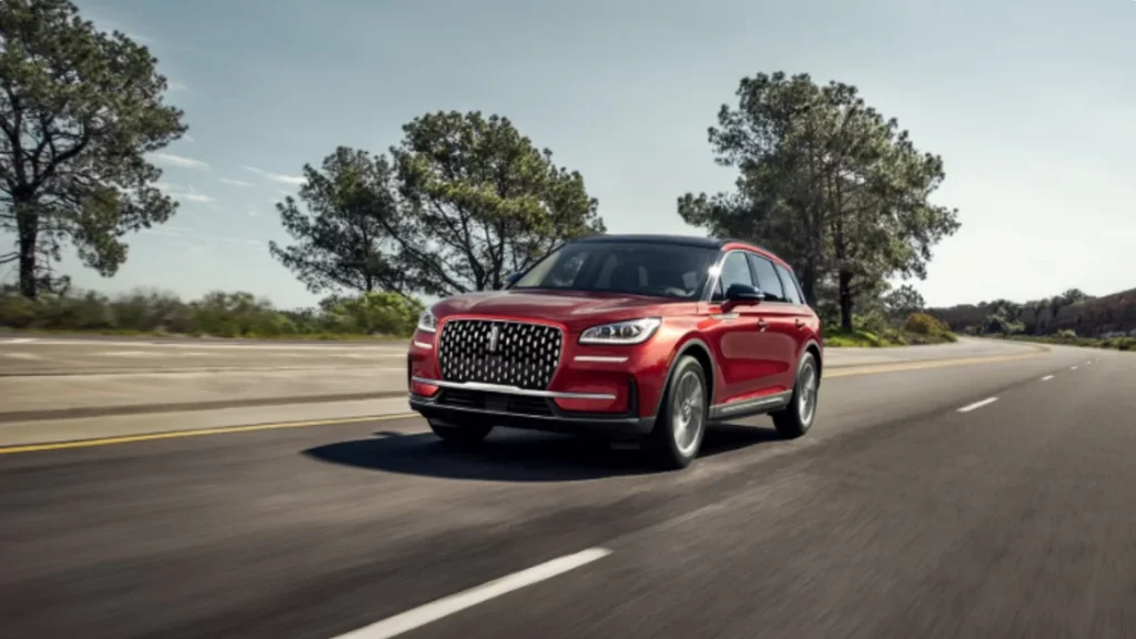 Red 2025 Lincoln Corsair® Grand Touring SUV driving along a sunlit highway with trees and blue sky background.
