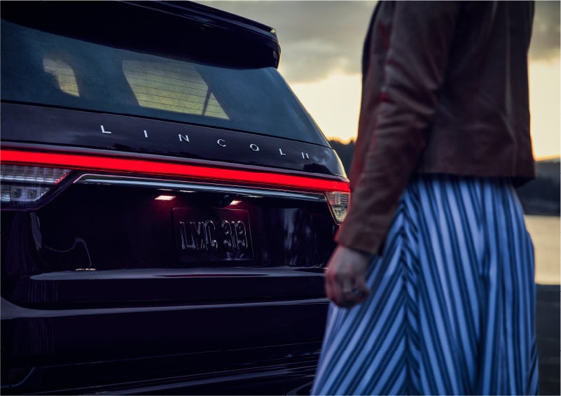 A person is shown near the rear of a 2023 Lincoln Aviator® SUV as the Lincoln Embrace illuminates the rear lights | Nick Mayer Lincoln Mayfield in Mayfield Heights OH