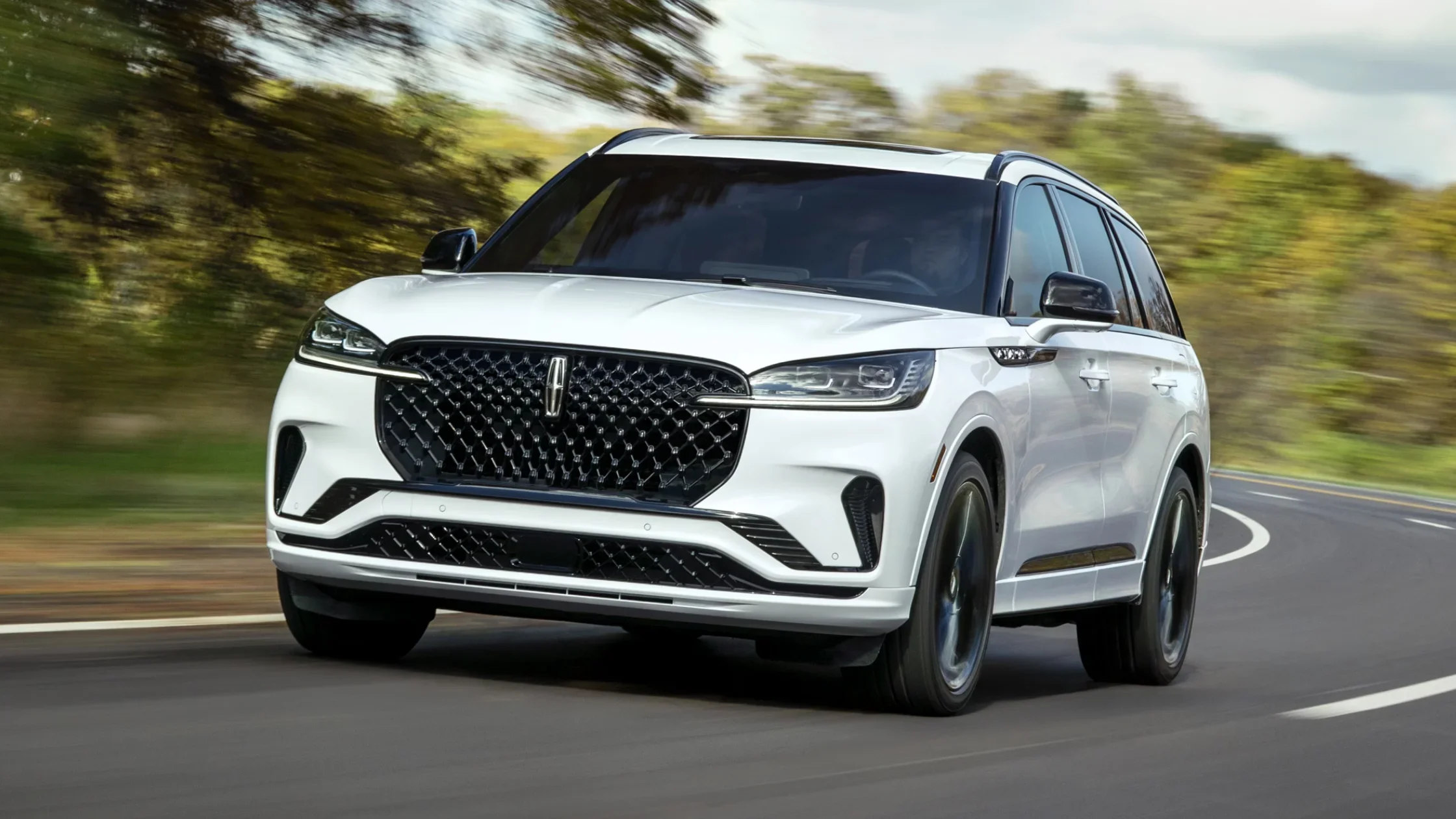 Front exterior view of a white 2025 Lincoln Aviator® Reserve SUV driving on a curved road with a black mesh grille and LED headlights.