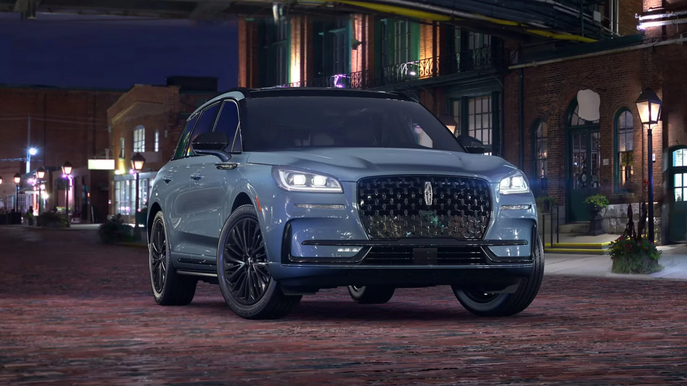 Blue 2025 Lincoln Corsair Grand Touring parked on cobblestone street at night with LED headlights and black grille.