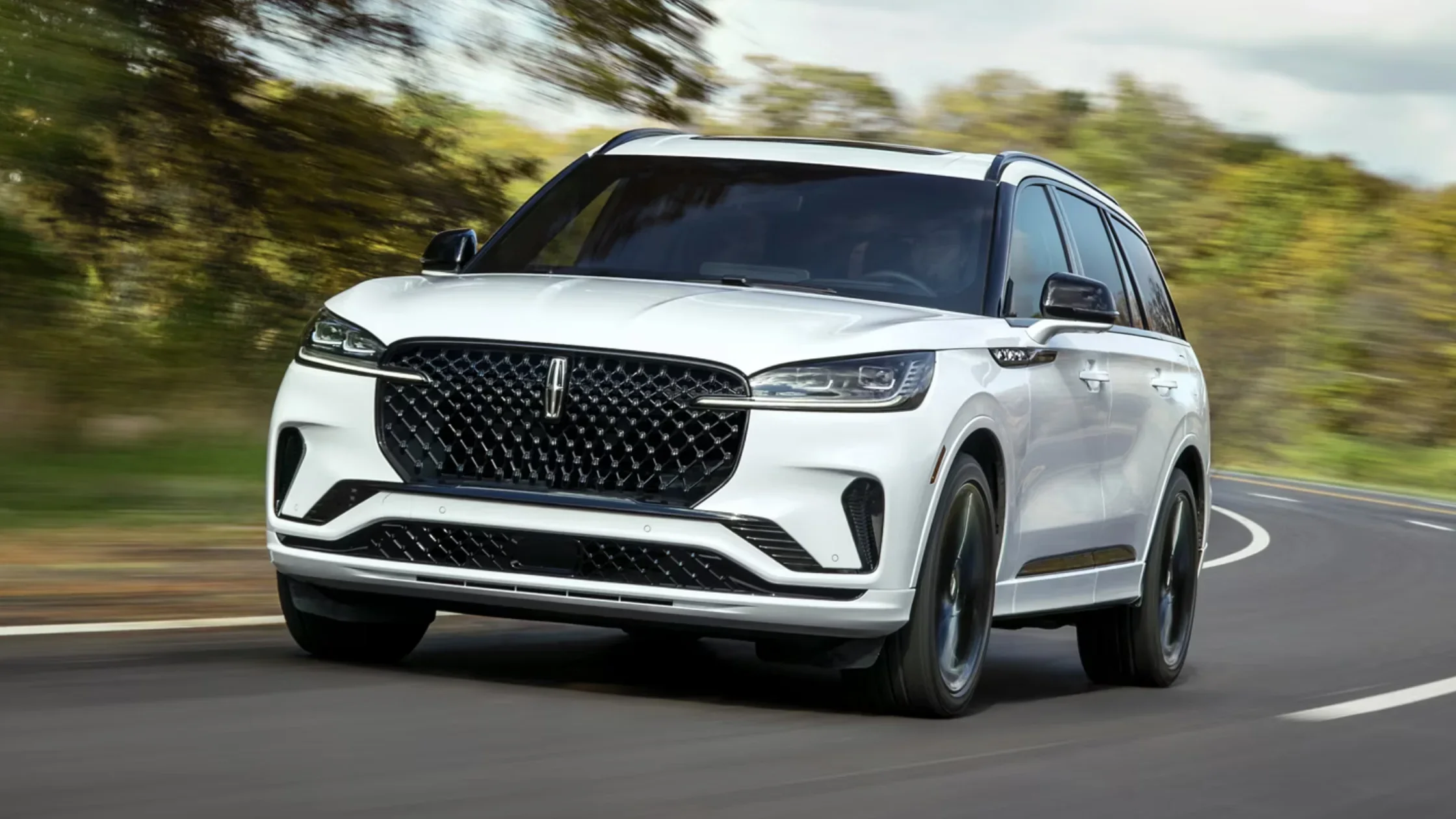 Front three-quarter view of 2025 Lincoln Aviator in white driving through a curved road, diamond mesh grille visible.