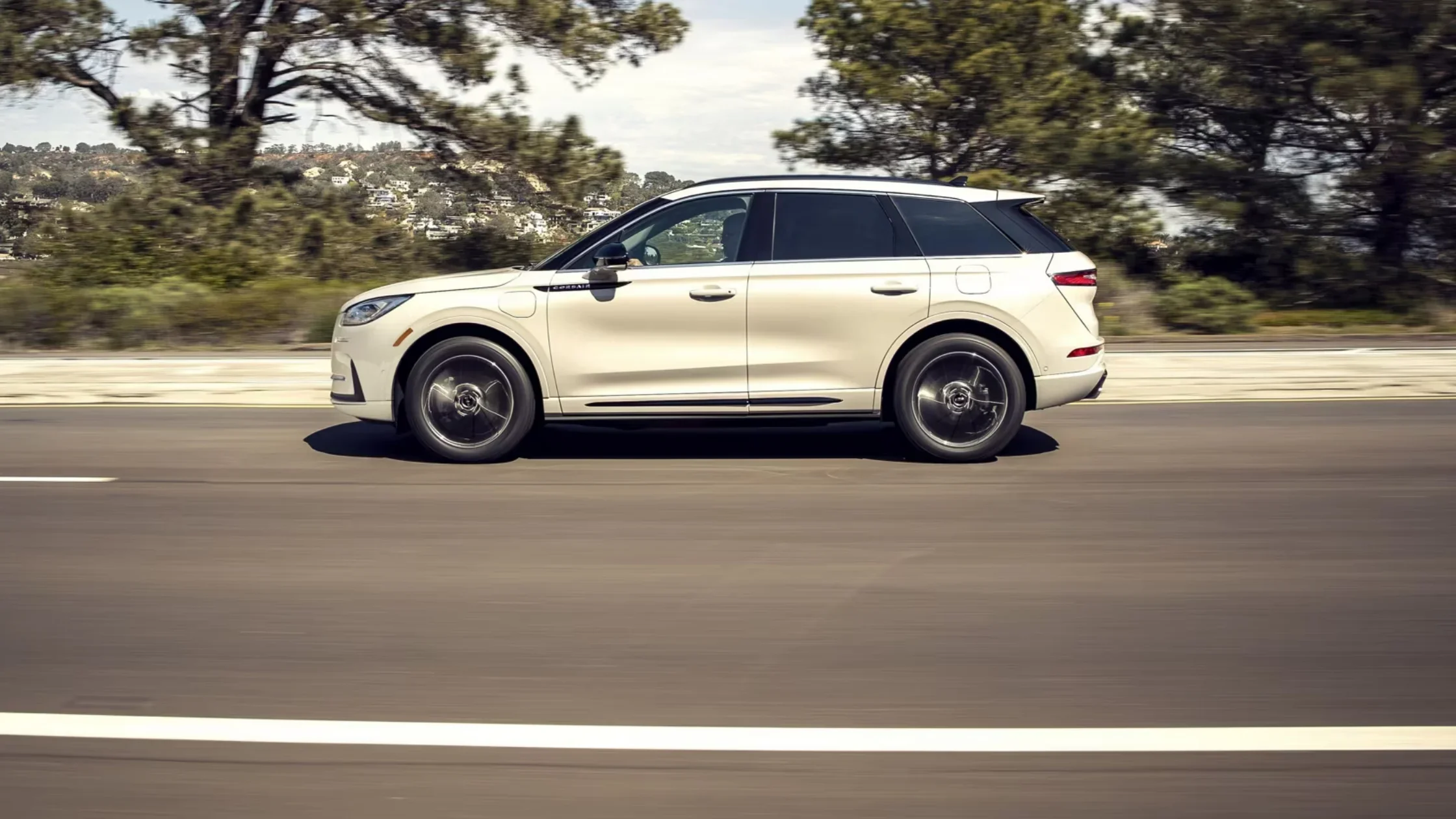 White 2025 Lincoln Corsair® side profile in motion on a highway flanked by trees and hills.