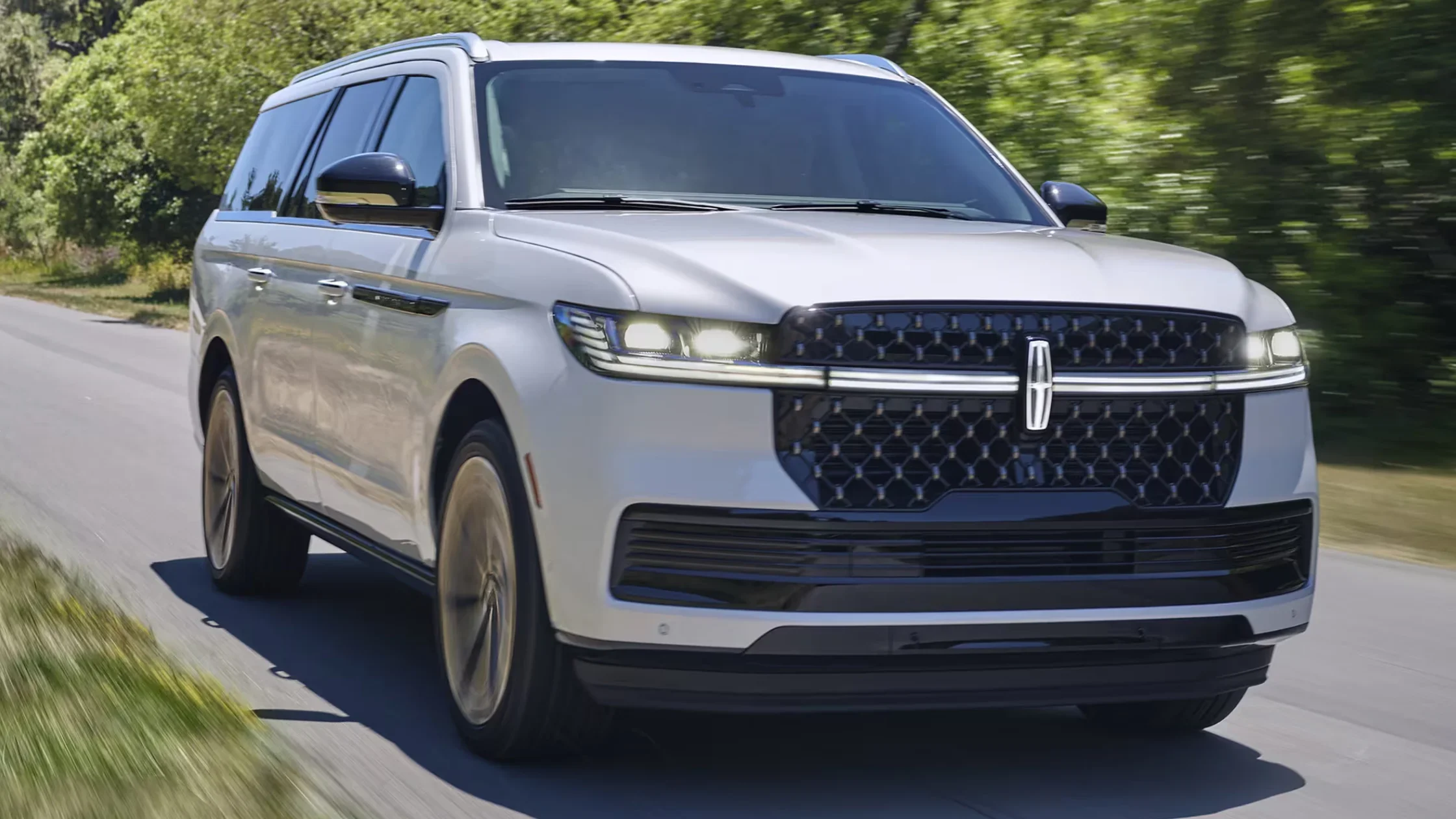 Front view of a white 2025 Lincoln Navigator® driving on a shaded road, showcasing its LED headlights and bold grille design.